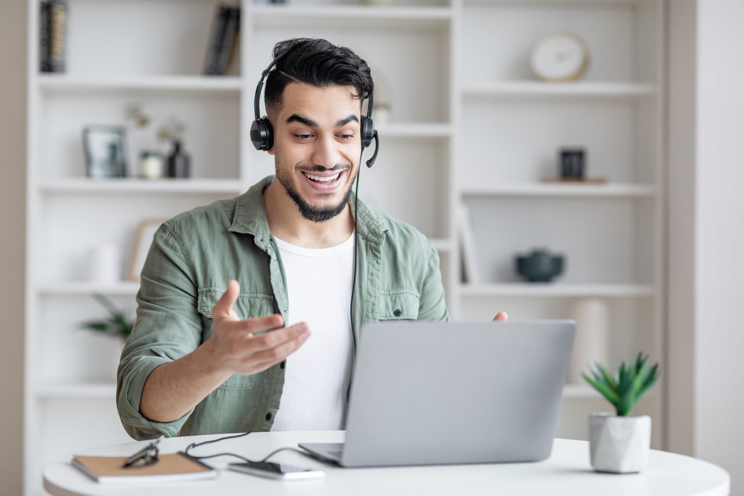 A smiling man with a beard, wearing a green shirt and headset, is gesturing during a video call on his laptop. White shelves with books and decorations are in the background.