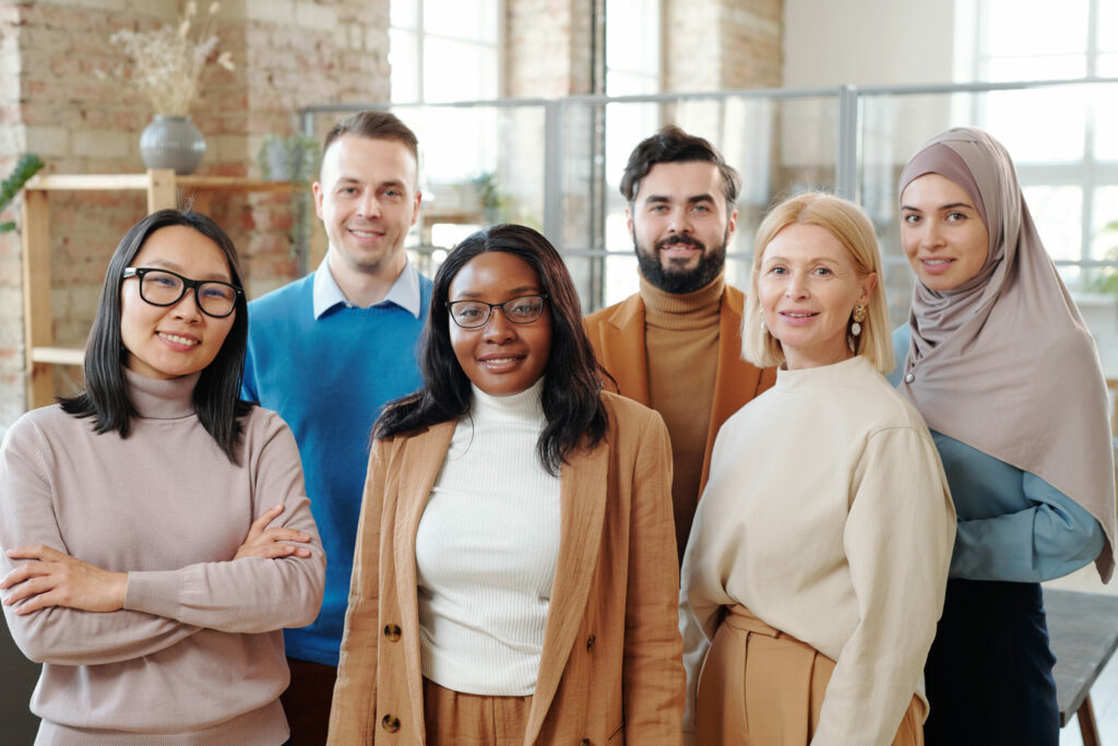 A diverse group of six professionals in business casual attire standing together in a modern office with exposed brick walls and large windows