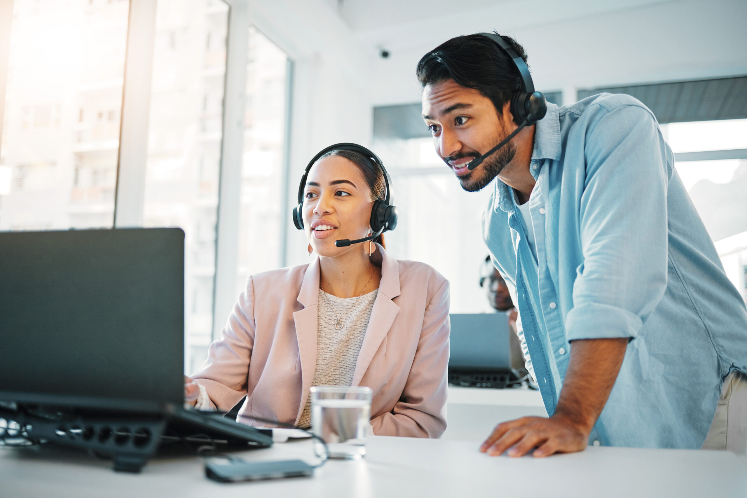 Two professional agents wearing headsets are looking at a laptop and discussing something in a bright, modern office.