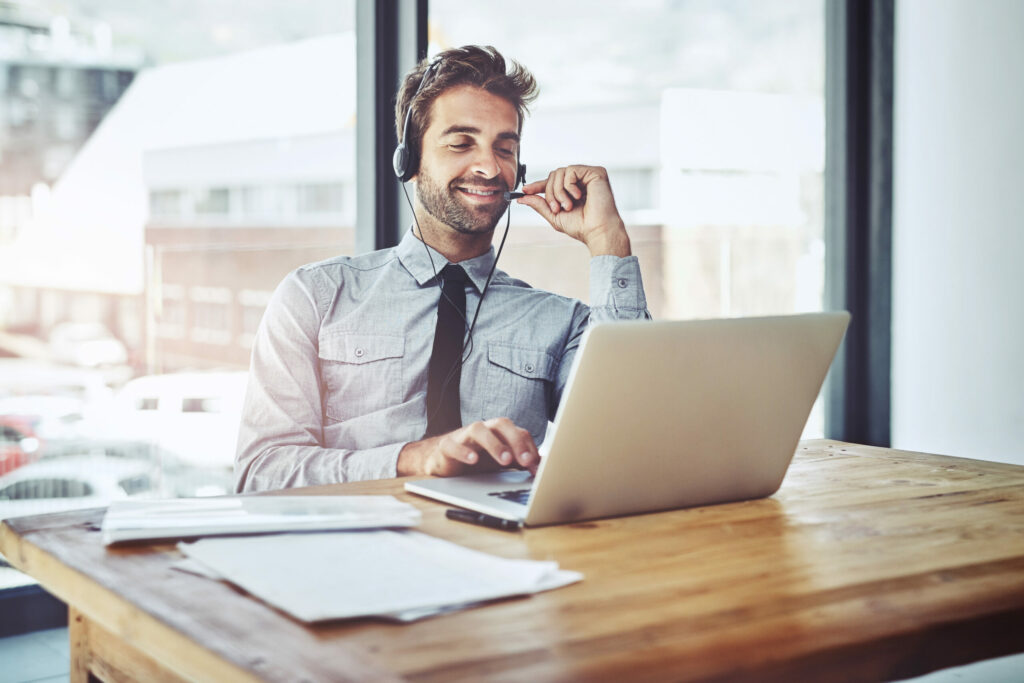 A professional man wearing a headset and working on a laptop, smiling while interacting with a customer. The scene is set in a modern office with large windows, suggesting a focus on customer experience (CX) and remote communication technology.