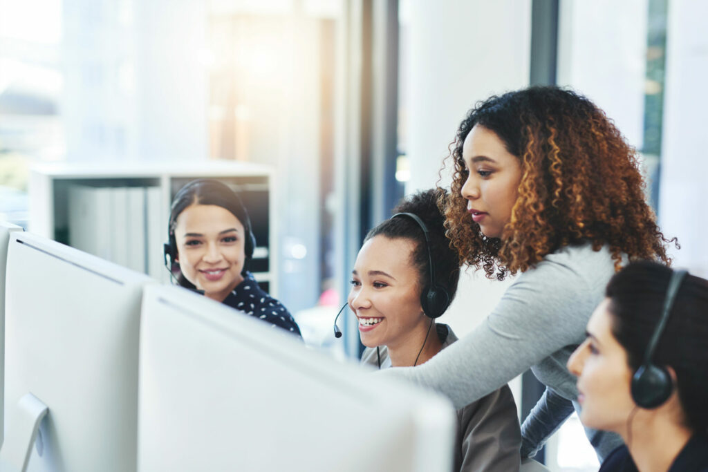 An office space with several desks and chairs arranged in a modern, open layout. Each desk has computer monitors, office supplies, and personal items, suggesting a busy and productive work environment. The lighting is bright, and the space is designed with a professional, contemporary aesthetic