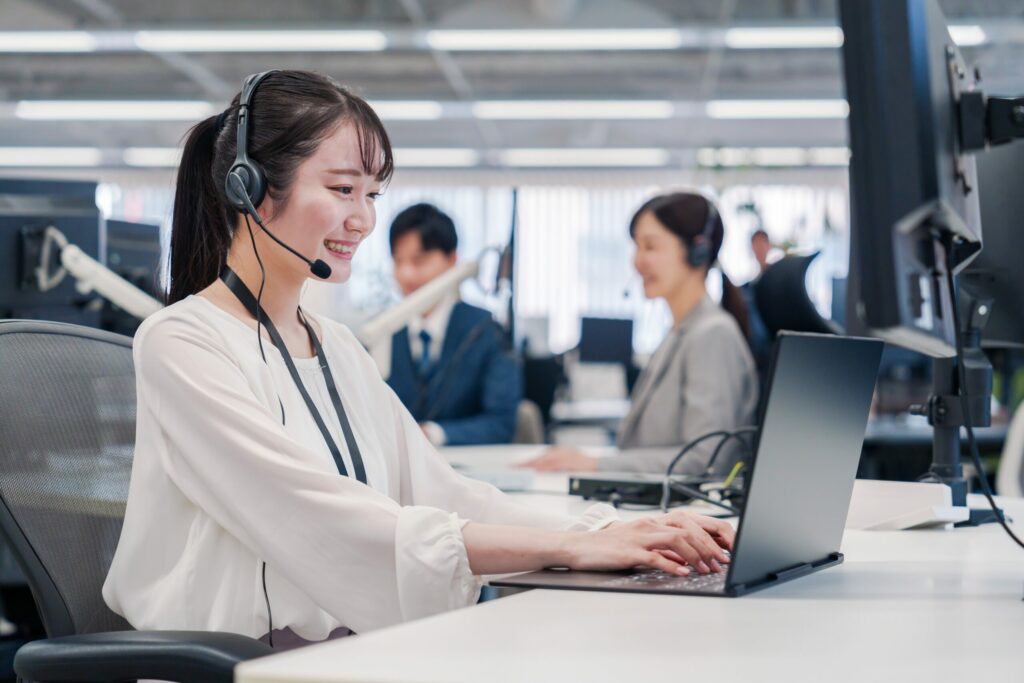 Smiling female call center agent wearing a headset and working on a laptop in a modern office. Other customer service representatives are visible in the background, engaged in their tasks
