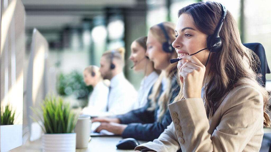 Smiling customer service representative wearing a headset, working in a modern contact center alongside colleagues, symbolizing innovation and excellence in customer experience outsourcing.