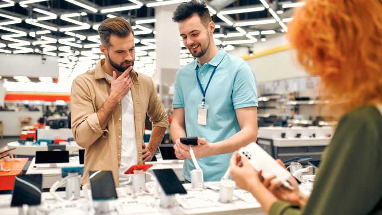 A man in a beige shirt is thoughtfully looking at a smartphone in his hand, guided by a store employee wearing a light blue polo shirt and a lanyard. The setting appears to be an electronics store with various smartphones displayed on a counter. Another person with orange hair, visible from behind, is also holding a smartphone, examining it. The store is brightly lit with modern, geometric lighting on the ceiling.