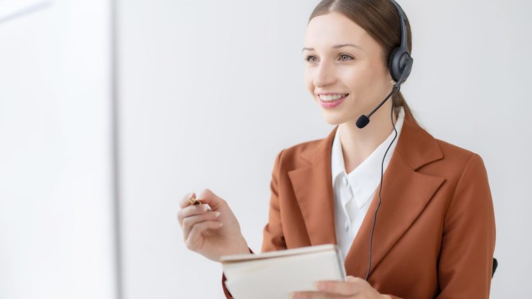 A customer service representative wearing a brown blazer and a white shirt is seated, holding a notepad and pen in her hands. She is smiling and wearing a headset with a microphone, suggesting she is engaged in a conversation or providing customer support. The background is simple and white, keeping the focus on her.