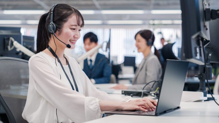Smiling female call center agent wearing a headset and working on a laptop in a modern office. Other customer service representatives are visible in the background, engaged in their tasks