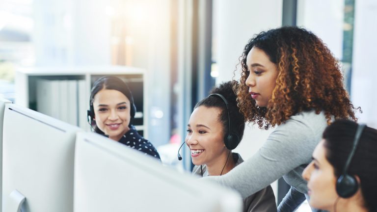 An office space with several desks and chairs arranged in a modern, open layout. Each desk has computer monitors, office supplies, and personal items, suggesting a busy and productive work environment. The lighting is bright, and the space is designed with a professional, contemporary aesthetic